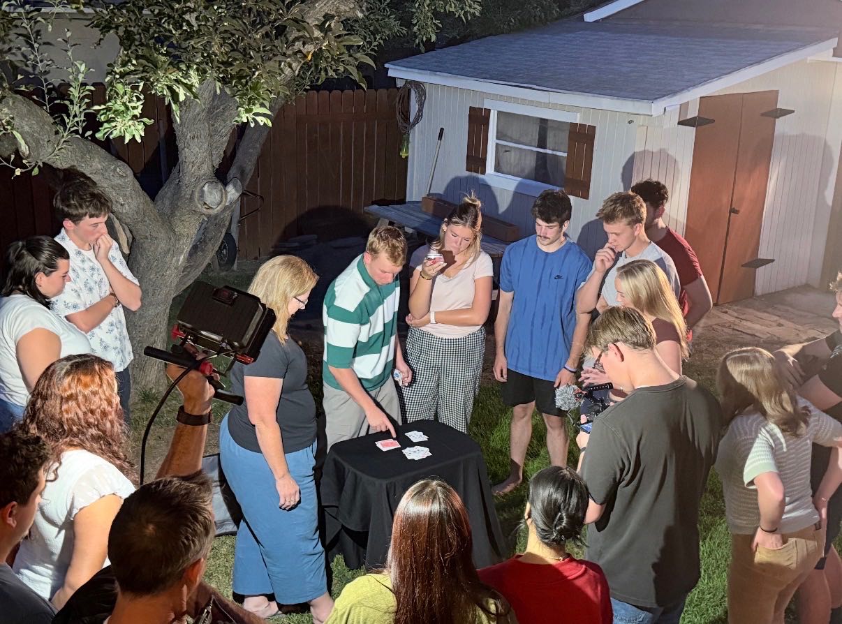 Ripley the Magician performing close-up card magic at a table for a circle of guests in a backyard at dusk.